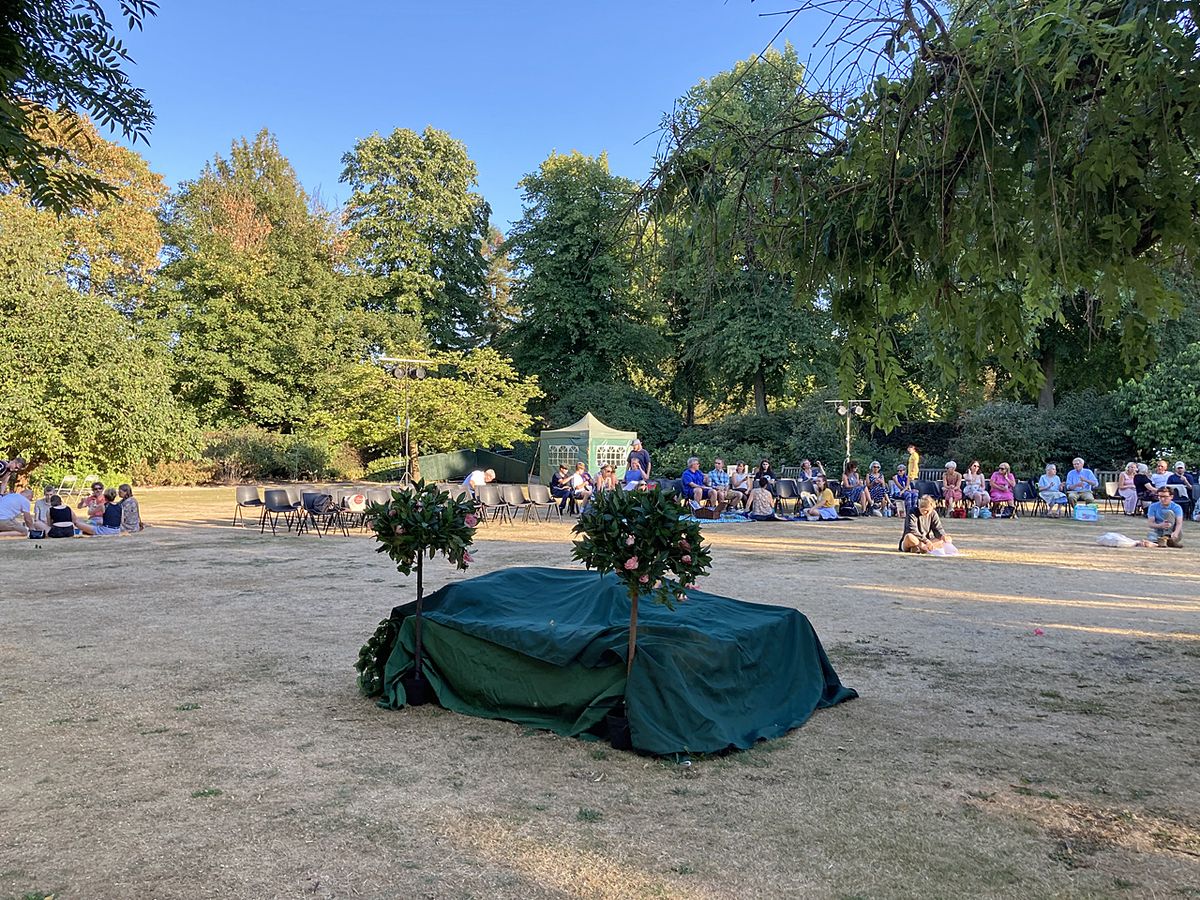 An audience on chairs and rugs in a college garden before an open-air Shakespeare performance